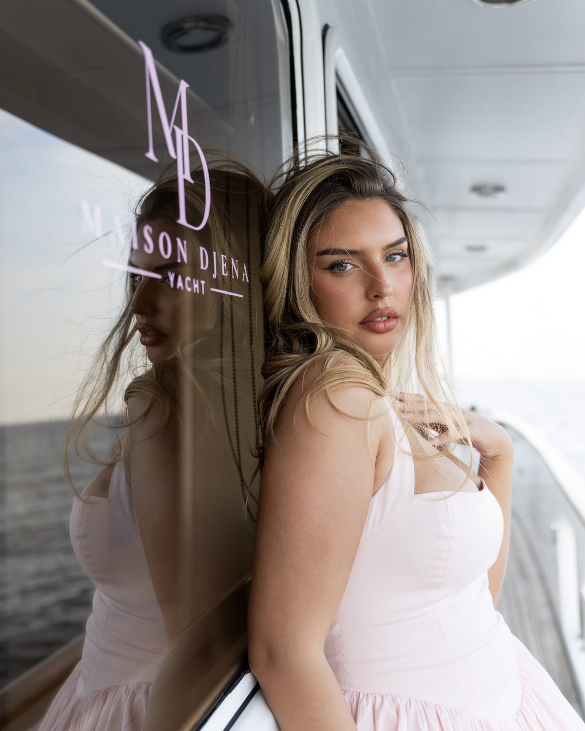 Djena adossée à la vitre du yacht, monogramme Maison Djena en reflet